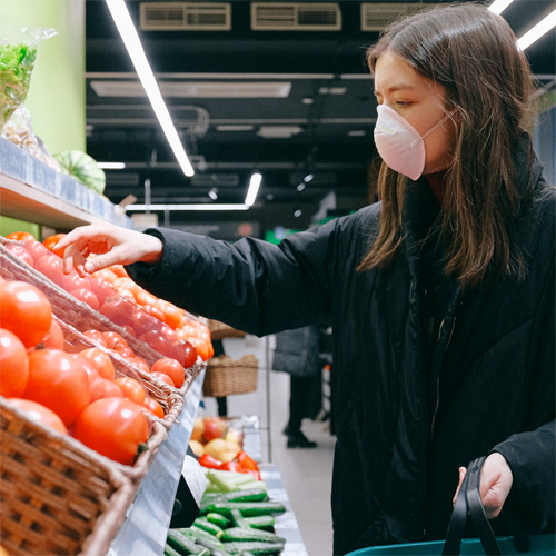 person in a mask picking out tomatoes from a store shelf