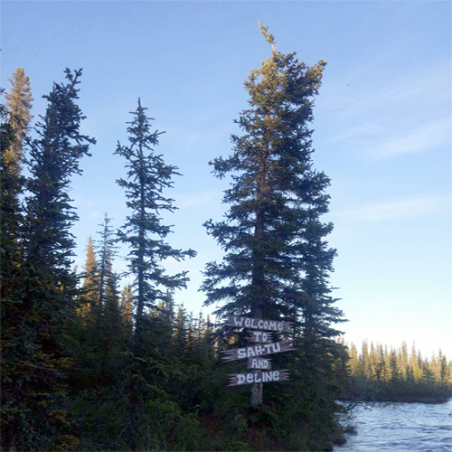 a tall evergreen tree with a hand-painted sign saying Welcome to Sah-tu and Délįnę